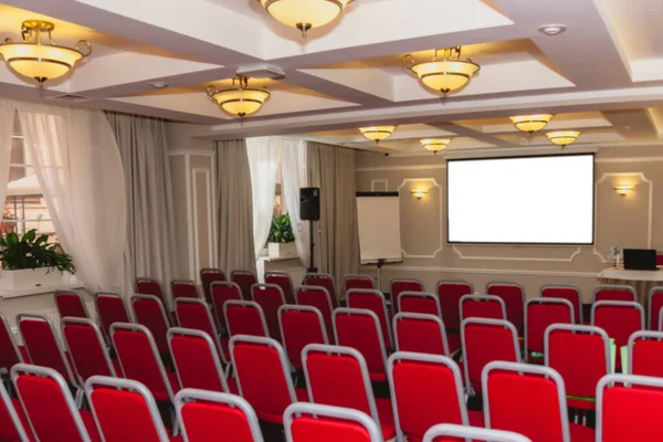 Conference room with red chairs, interior lighting, and empty white insulated screen. Business meetings, conferences, trainings, used as template for elegant design. Interior of the conference hall