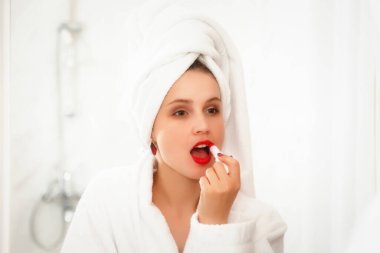 Kadın duştan sonra otel banyosunda dudaklarını boyuyor. Aynayla ilgili sabahlık giymiş Slav görünümlü güzel bir kadın. Mutlu bayan. Sağlıklı yüz derisi. Sevimli kız yüzünü yıkadı ve duygularını gösterdi.