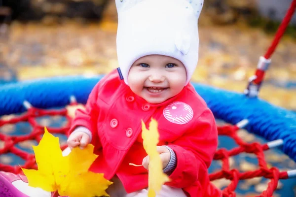 孩子们骑着黄色的秋叶荡秋千 可爱的蓝眼睛的一岁小女孩 身穿红衣 头戴白帽 迷人的婴儿在特写中表现出感情 健康儿童和养育子女的概念 — 图库照片