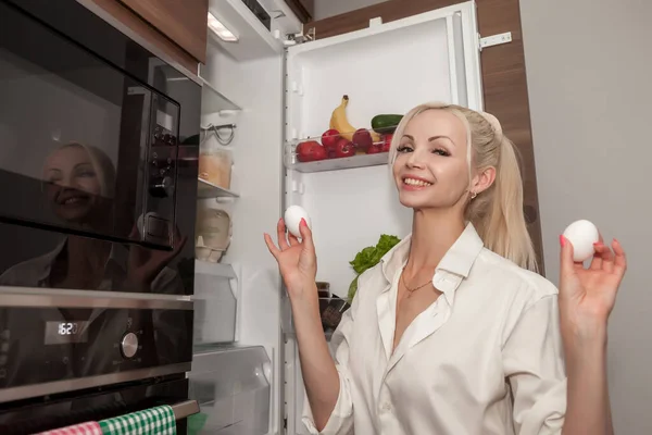 Woman at an open refrigerator with chicken eggs in hands. Modern attractive girl in kitchen, opens refrigerator door and takes food. Young cheerful female takes eggs from freezer and smiles at camera