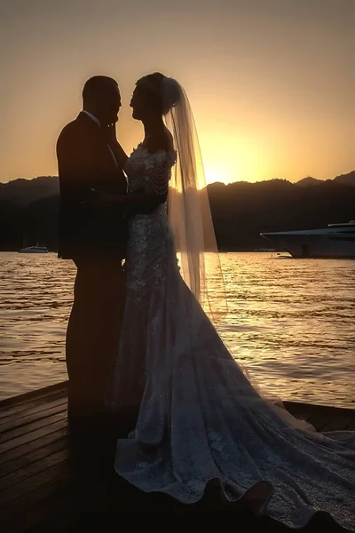 結婚式の日の日没時に海や海の新郎新婦 山の後ろに行く太陽の背景に結婚式のドレス ドレスやベール で花嫁のシルエット 結婚式の日の終わり — ストック写真