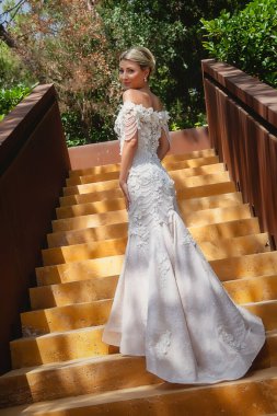 Gelinlik giymiş ince bir kadın güneşli bir günde merdivene tırmanır. Gelin, kameraya bak. Güzel bir düğün sabahı. Çapraz görüntü. Harika bir kız. Kadınlar duygularını gösterir. Mavi gözlü sarışın.