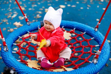 Çocuk sarı sonbahar yapraklarıyla salıncakta sallanıyor. Kırmızı ceketli, beyaz şapkalı, mavi gözlü, şirin bir kız. Sevimli bebek, yakın planda duygularını gösterir. Sağlıklı çocuk ve ebeveynlik kavramı