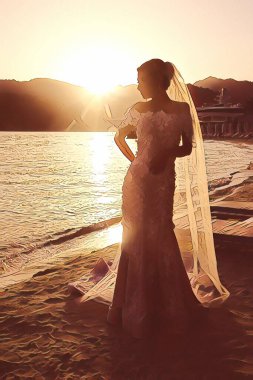 Düğün günü gün batımında okyanusta ya da denizde gelin. Gelinin gelinlik ve duvak giymiş silueti, dağların arkasına giden güneşin arka planına karşı. Düğün gününün sonu.