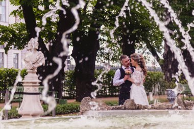 Gelin ve damat, çeşmenin arkasında akan suyla Park 'ta dururlar. Güneşli düğün gününde gelinlik giyen yeni evliler. Sokakta harika manzaralı bir çift. Yeni evliler birlikte mutlular.