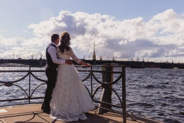 Gelin ve damat, nehir ve katedralin arka planında birbirlerine sarılıyorlar. Güneşli düğün gününde gelinlik giyen yeni evliler. İnanılmaz manzaralı bir çift. Yeni evliler birlikte mutlular.