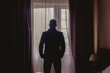 Otel odasının penceresinde duran zarif takım elbiseli genç adamın portresi. Erkekler için düğün sabahı. İç odada damat kontur ışığı altında. Dikiz aynası. Telif Hakkı Boşluğu