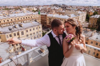 Gelin ve damat, panoramik manzarası olan eski kasabayı çatıda kucaklıyor. Güneşli düğün gününde gelinlik giyen yeni evliler. Çatıda muhteşem manzaralı bir çift. Şehrin arka planında aşık yeni evliler