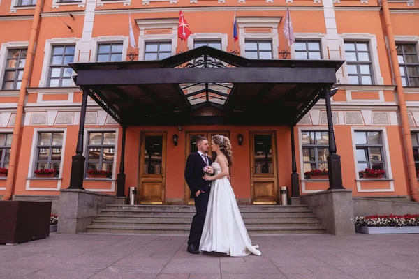 Bride and groom stand embracing main entrance to building with an old facade. Newlyweds in wedding dresses on Sunny wedding day. Couple on street in amazing view. Newlyweds in love happy together