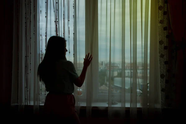 Female silhouette against window. Woman stands and looks out the window at city on high floor. Girl in background light. Concept of living in big city or loneliness at home. Copyright space