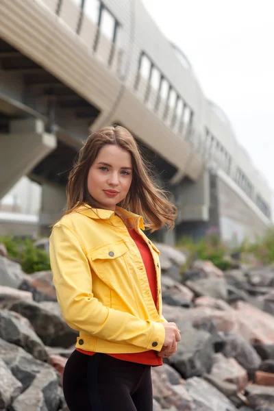 身材魁梧 性感的年轻女子站在海滩上的石头上 穿过市中心 街上穿着黄色夹克和黑色长裤的可爱女人的画像 — 图库照片
