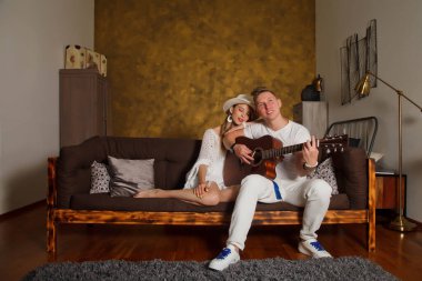 Odanın içinde gitarlı harika genç bir çift. Kanepede gitar çalan tatlı bir kadın ve erkek. Evde gitar çalma ya da öğrenme kavramı. Site, bayrak veya logo için telif hakkı alanı