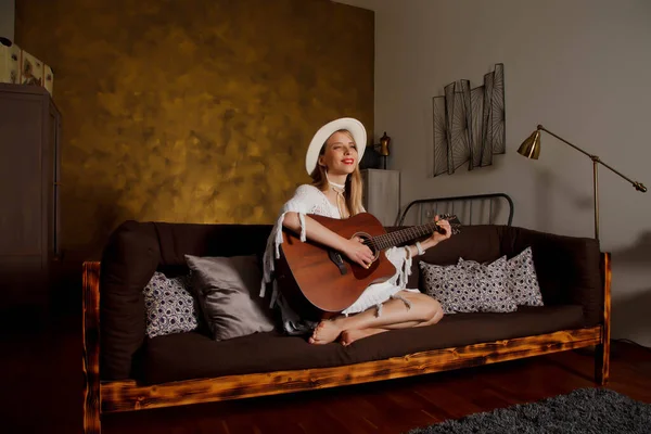 Beautiful young woman with guitar in interior of room. Cute woman on couch with guitar in her hands. Concept of home learning or playing guitar at home. Copyright space for site or banner or logo
