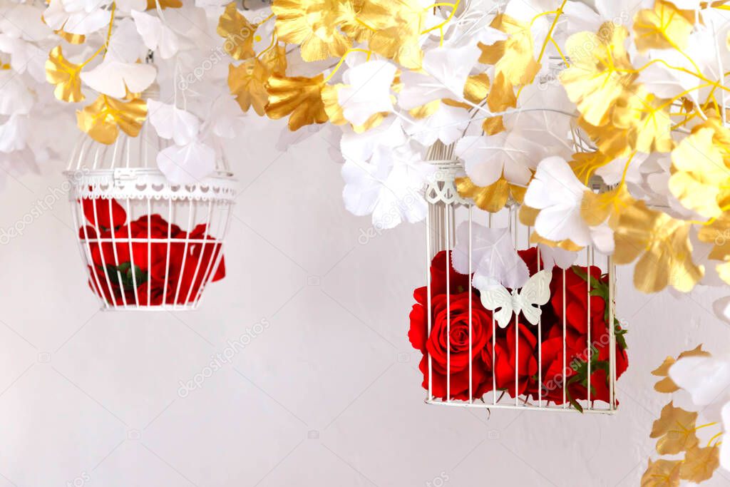 Decorative bird cages on white wall background with red roses. Amazing wedding decor in interior. Flowers in birdcage for decorating room or hall for Valentine's day. Text or logo on site. Copy space