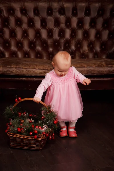 Cute one-year-old girl in pink dress on background of sofa with basket Christmas decorated toys. Child in decorations with gift presents. Concept of cozy home Happy New Year celebration. Copy space