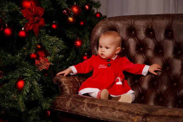 Cute one-year-old girl in Santa Claus costume on sofa on background of Christmas tree decorated toys. Child in decorations with gift boxes. Concept of cozy home Happy New Year celebration. Copy space