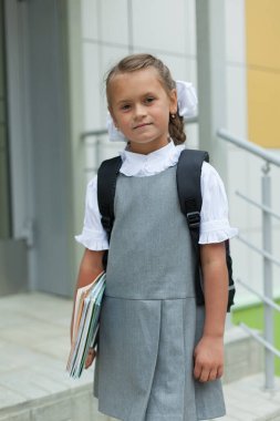 Not defterleri ve sırt çantası olan küçük bir kız okulun yanında duruyor. Birinci sınıf öğrencisi. Bilgi Günü