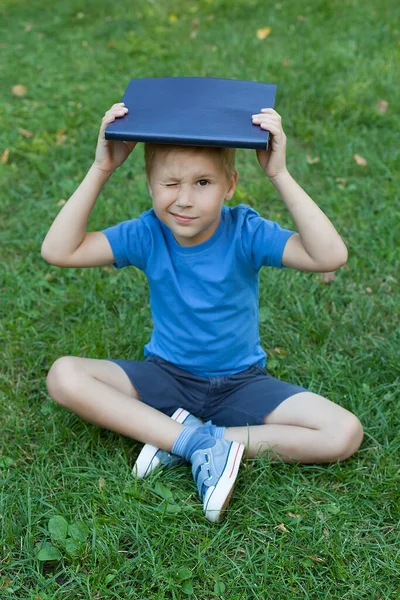 Um menino pequeno no parque está e prende um livro em sua cabeça. Ler um livro. — Fotografia de Stock