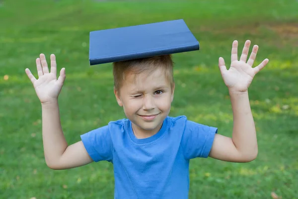 A cute boy sits on the grass in a Park and holds a book on his head, winking an eye. He raised his hands. — Stock Photo, Image