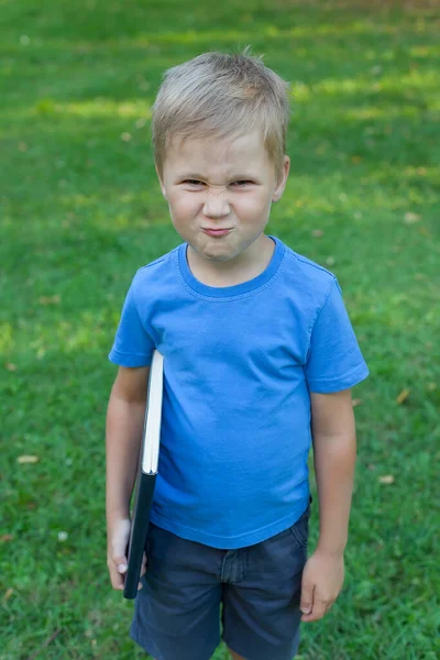 Um menino pequeno no parque está e prende um livro em suas mãos. ele enruga o nariz e sorri. — Fotografia de Stock