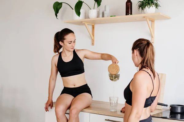 Two sports girls spend time in the bright kitchen. Women in sports shorts and tops are sitting on the kitchen furniture and drinking lemonade. Communication girls at home. Healthy eating