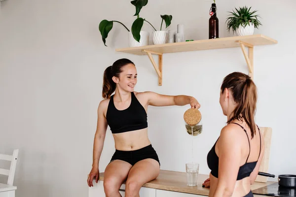 Two sports girls spend time in the bright kitchen. Women in sports shorts and tops are sitting on the kitchen furniture and drinking lemonade. Communication girls at home. Healthy eating