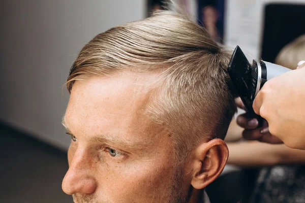 A blond hair man is hair cutting with barber in the barbershop. Haircut with an electric razor. Self-care. Close-up haircut