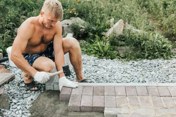 A blond sporty man is laying paving slabs. Arrangement of the yard. Outdoor work with a mallet and paving stones. selective focus.