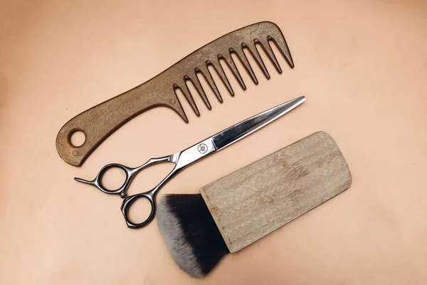 Wooden comb, scissors and paintbrush on a beige background. Hairdressing tools.