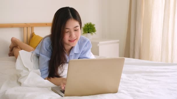 年轻的亚洲女人在家里靠电脑笔记本电脑在床上工作 开心地笑着看着相机 — 图库视频影像
