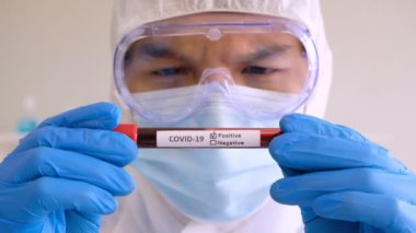 Bilim adamları hijyen maskesi takıyor ve laboratuarda analiz için pozitif Cauvid-19 virüsü etiketli kan testi tüpü alıyor..