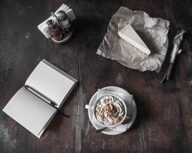 Karanlık fotoğraf. Bir defter ve peynirli kekin yanında krem şantili cappuccino. Samimi kahve dükkanı, seçici odaklanma.