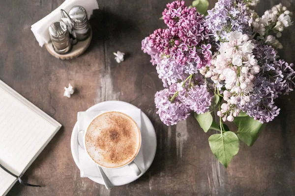 Kahve dükkanında, sehpanın üstünde. Bir leylak ve bir not defterinin yanında tarçınlı cappuccino, seçici odak