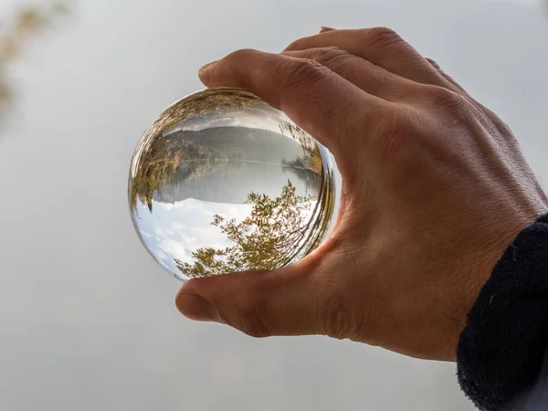 Closeup of a glass ball held up by a human hand reflecting the wooded area inside the sphere. Forest lens ball