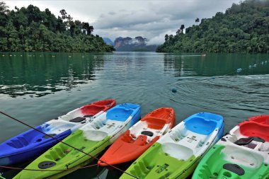 Gölün sakin, parlak yüzeyinde parlak, renkli kanolar vardır. Uzakta sıradışı şekilli kayalık adalar, tropikal bir orman. Alçak bulutlar ve sis. Tayland., Cheo Lan.