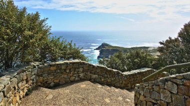 Etrafı çalılarla çevrili taş bir merdiven dağdan iner. Atlantik Okyanusu ve Ümit Burnu manzarası. Güneşli bir gün, gökyüzündeki bulutlar. GÜNEY Afrika.                               