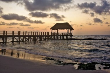 Karayip Denizi üzerinde gizemli bir gün doğumu. Küçük dalgalar pürüzsüz kumsalda yuvarlanıyor. Suyun üzerinde ahşap bir patika ve bir kubbe var. Gökyüzü turuncu ve pembe, leylak bulutları. Meksika peri masalı.