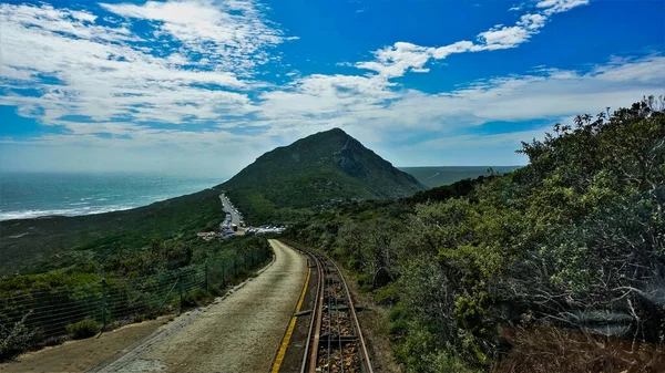 山中的路 有车也有车 有车也有车 路边的绿色植物 这条公路藏在一座小山后面 大西洋可见 天空明亮 南非开普敦 通往开普敦的路 — 图库照片