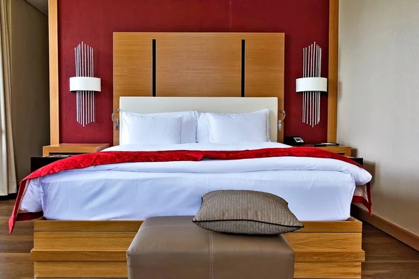 The interior of the hotel. Double bed with white linen and red bedspread. Wooden panel at the head, lamps. At the feet of the couch, a pillow. Beige walls.
