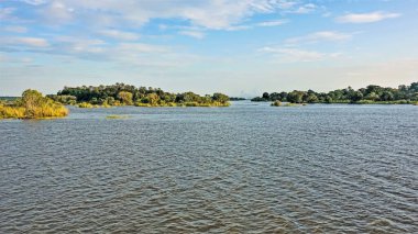 Yaz günü. Sakin nehir adalarında tropikal bitki örtüsüyle. Victoria Şelalesi üzerinde sisli bir sis görünüyor. Mavi gökyüzü ve ışık bulutları. Zimbabwe. Zambezi nehri.