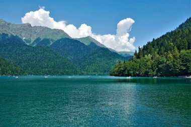 Alp Gölü Ritsa. Tekneler pürüzsüz turkuaz suda yüzerler. Dağ yamaçları sık ormanlarla kaplıdır. Gök kubbede resim gibi bulutlar var. Abhazya.