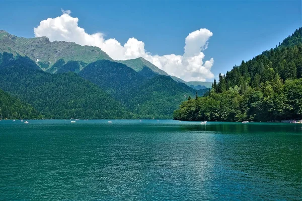 Alp Gölü Ritsa. Tekneler pürüzsüz turkuaz suda yüzerler. Dağ yamaçları sık ormanlarla kaplıdır. Gök kubbede resim gibi bulutlar var. Abhazya.