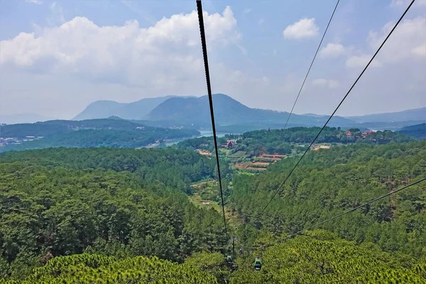 Cable car cables run over the forest. In the foreground are dense thickets of coniferous trees. Lake in the distance. Beautiful mountains against the blue cloudy sky. Vietnam. Da lat.
