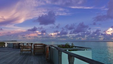 Okyanusun ortasında tahta bir güvertede masalar ve sandalyeler var. Sakin turkuaz su. Akşam gökyüzünde pembe ve eflatun bulutlar var. Maldivlerde gün batımı.