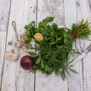 Beyaz ahşap bir masanın üzerinde kırmızı bir iple bağlanmış bir demet maydanoz ve dereotu uzanır. Yanında dilimlenmiş limon, sarımsak, kırmızı soğan ve bıçak var. Bir çay kaşığında ve masada baharatlı gri tuz var..