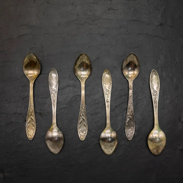 Set of six tiny cupronickel coffee spoons. Decorated with floral ornaments, covered with patina. The beginning of the 20th century. The background is black stone tiles.