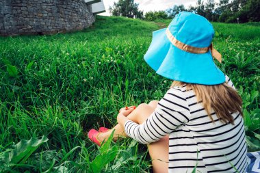 Yazın parkta yeşil çimlerin üzerinde mavi şapkalı bir kız oturuyor.