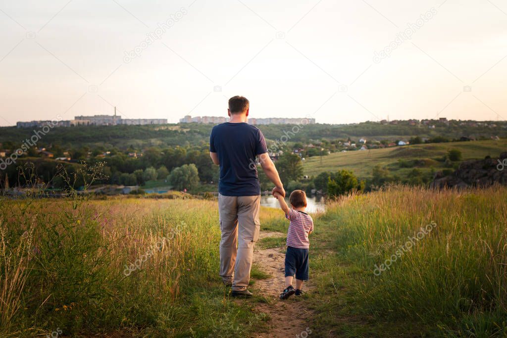 Dad and son walk in a yellow summer field in the evening at sunset and eat candy on a stick. Father's Day, love in the family, the role of the father in raising the child