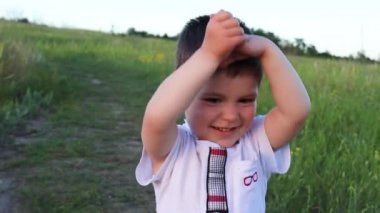 Ağır çekim. 3 yaşında beyaz tişörtlü bir çocuk ellerini kaldırıp gülümsüyor. Sahada çocukluk, mutluluk. Ailelere, anne ve babalara bir gülücük..