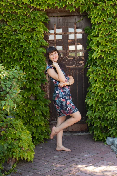 Vacker flicka 25 år brunett står barfota nära en trä magisk dörr, omgiven av gröna växter. — Stockfoto